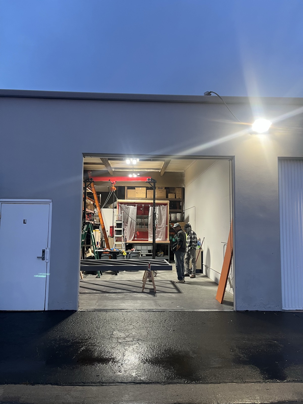 The test bay at dusk, seen from outside: gantry crane, curtained pressure chamber, and technicians reviewing the day's setup