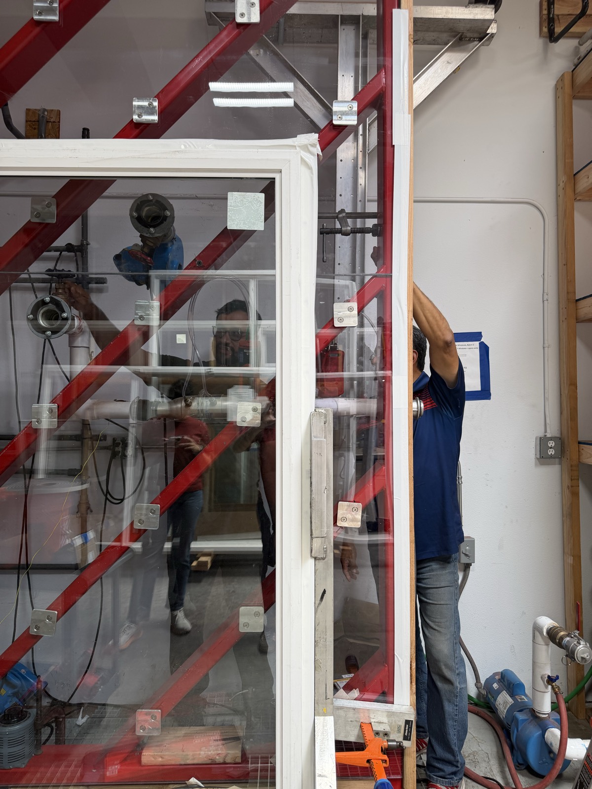 Window specimen mounted in a red-braced pressure test chamber with spray and pressure instrumentation