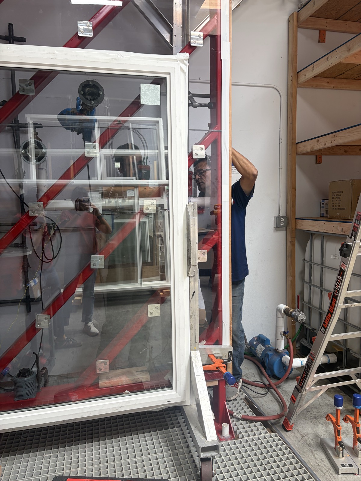 Jimmy Cruz positioning a single-hung window specimen in the red pressure test wall, with water supply pump and hoses plumbed at right
